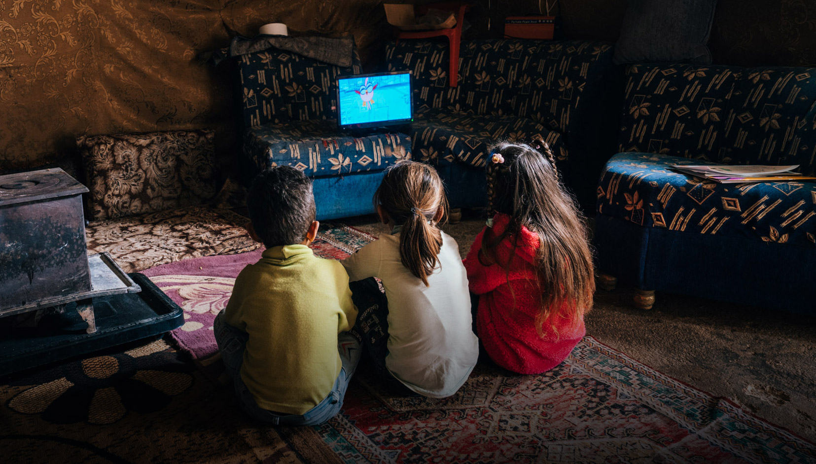 three children sit on the floor and watch a tablet propped up on a chair in front of them