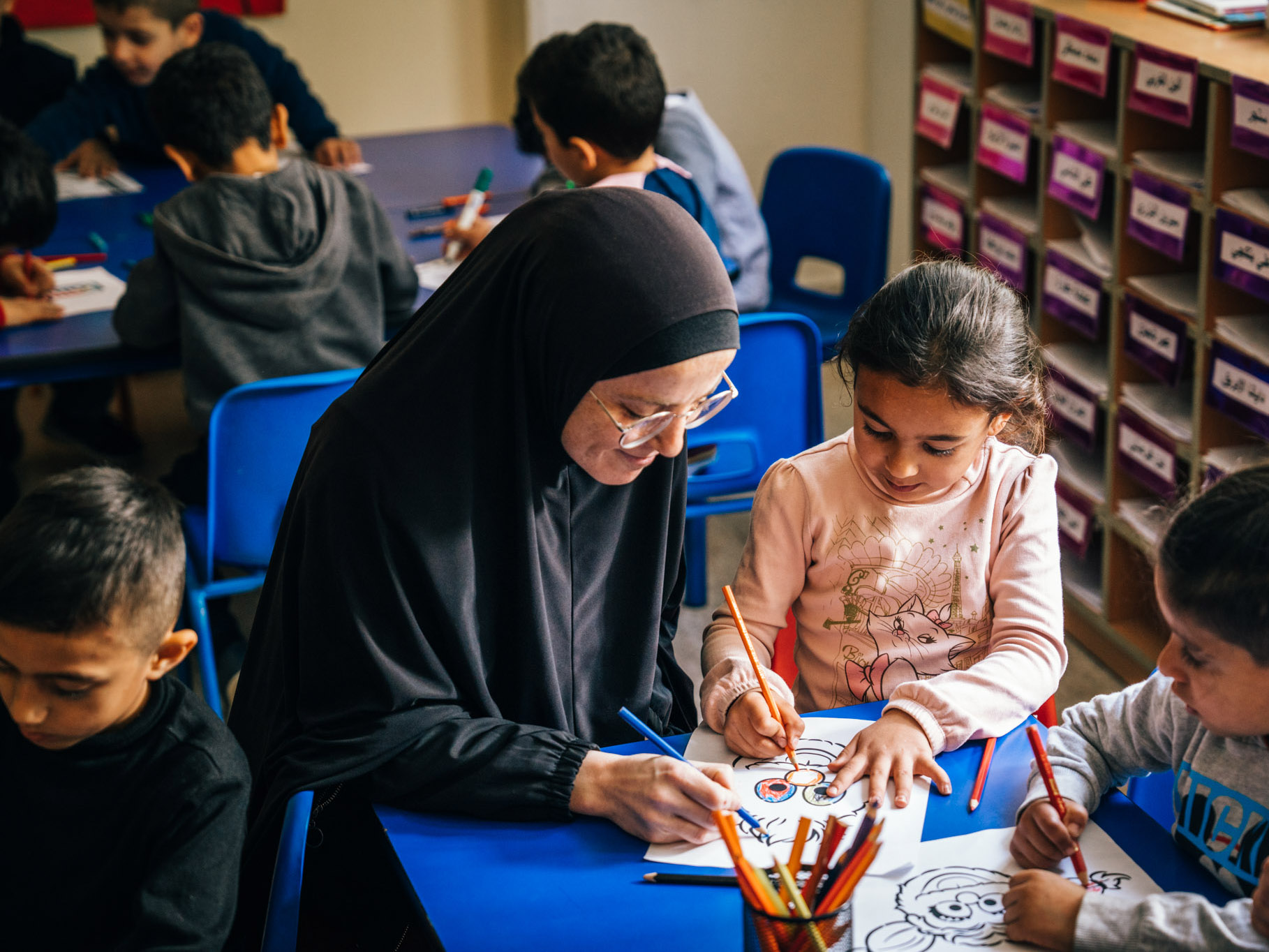 a teacher sitting at a table to help a preschool age girl with her coloring